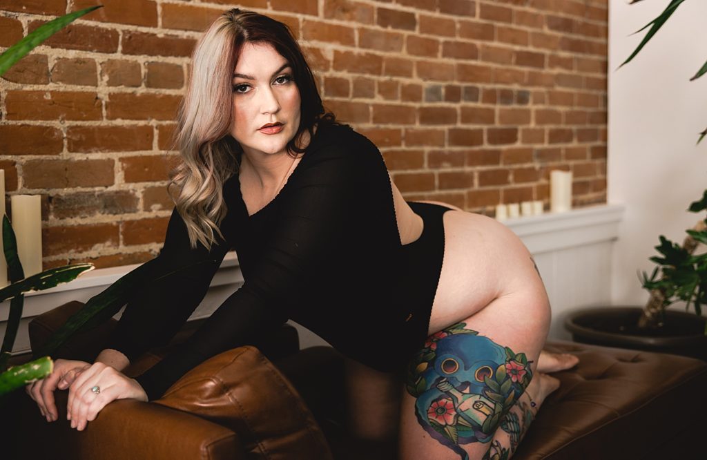 Woman with two tone hair and tattoos kneeling on a leather couch for her sexy photo shoot with Kansas City Photographer, WMNKND. 